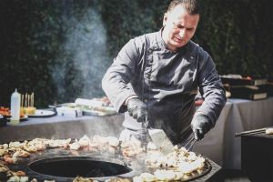 Ein Mann grillt auf einem großen Grill. Er hat eine typische Kochschürze an, die schwarz ist. Im Hintergrund sieht man ein Buffet aufgebaut.