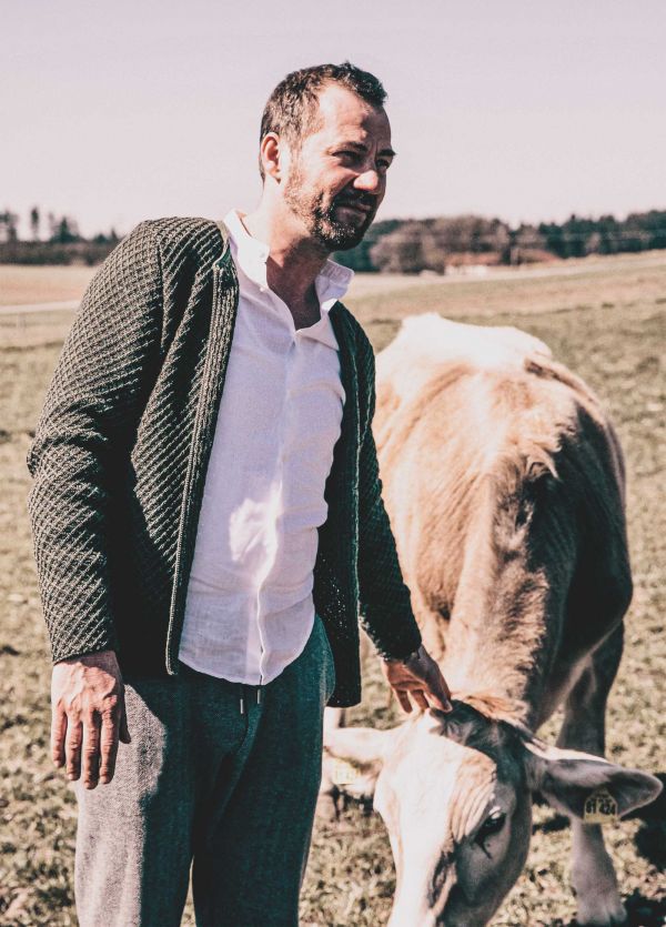 Alexander Tremmel steht auf einem Feld und schaut nach rechts. Dabei streichelt er ein Rind, welches gerade Gras frisst.