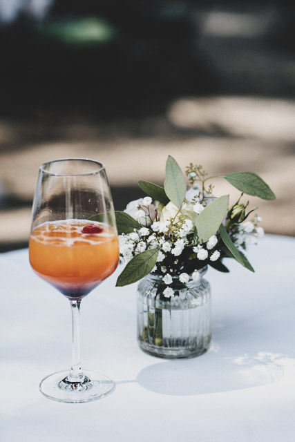 Auf einem Tisch mit weißem Tischgedeck steht ein Glas mit Alkohol und Blumendekoration.